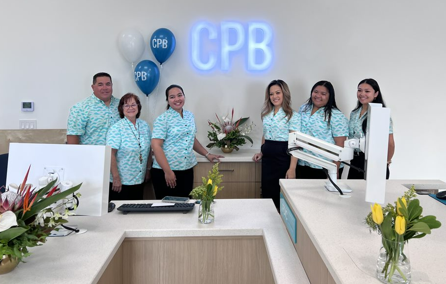 A group of six Central Pacific Bank employees pose behind a customer service desk inside a newly renovated branch. CPB-branded balloons, floral arrangements, and an illuminated CPB logo are displayed in the bright, modern banking space.