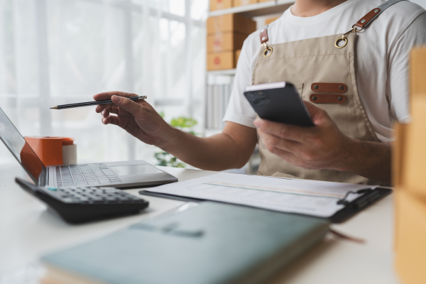 A small business owner sits at a desk holding a smartphone and pen while reviewing documents beside a laptop. A calculator, clipboard, and office supplies are visible, highlighting financial management and business operations.