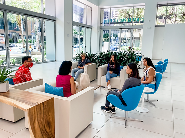 A group of people sit in a bright, modern bank lobby participating in a small discussion or meeting. Floor-to-ceiling windows, indoor plants, and blue lounge chairs create an open and welcoming setting.