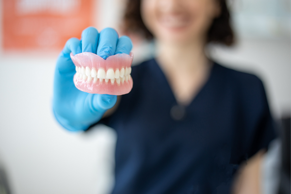 Person wearing a blue medical glove holding a model of human teeth, with the person smiling in the background.