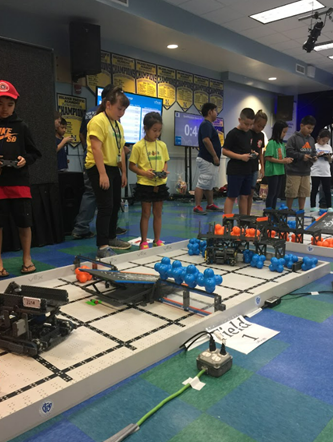 A group of students stand around a robotics competition field, operating robots with handheld controllers during a STEM event. The game field contains blue and orange scoring objects, while teams watch and compete in the background.
