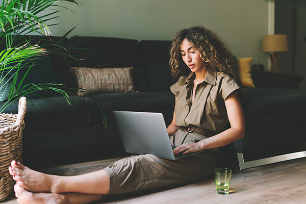 Person sitting on the floor of a living room using a laptop, with a sofa, houseplants, and a glass of water nearby.