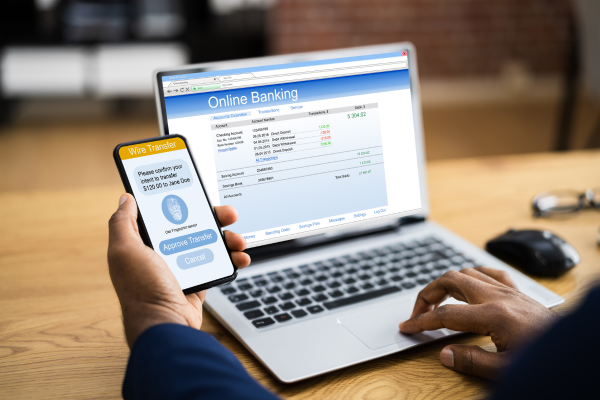 Person using online banking on a laptop while holding a smartphone displaying a wire transfer approval request, representing digital banking and payment security.