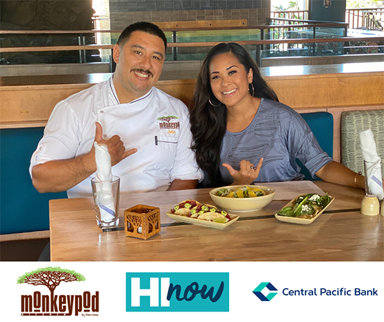 A promotional photo featuring two people seated at a restaurant table with several plated dishes, making shaka hand gestures. Monkeypod, HI Now, and Central Pacific Bank logos appear along the bottom of the image.