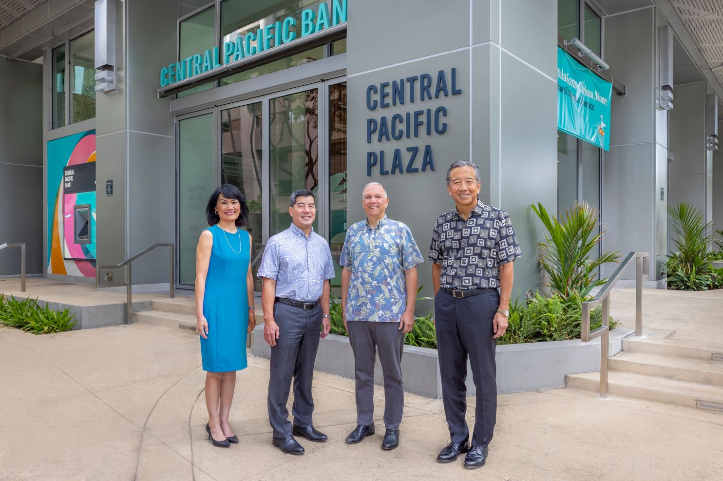 Four people stand outside the entrance of Central Pacific Plaza, posing in front of Central Pacific Bank signage and the building’s glass doors. Tropical landscaping and the modern exterior of the downtown bank building are visible in the background.