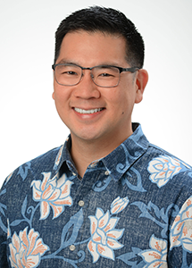 A professional headshot of a person wearing glasses and a blue floral aloha shirt, photographed against a light gray background.