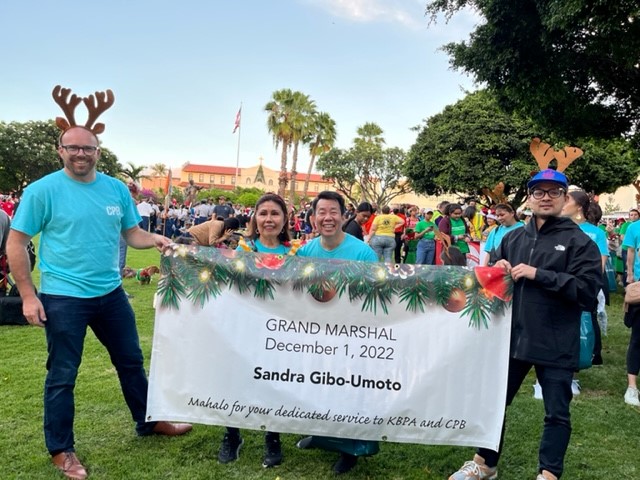 Four people stand in a park holding a banner recognizing Sandra Gibo-Umoto as Grand Marshal on December 1, 2022. The group poses among a large holiday crowd, with some participants wearing reindeer antlers and festive decorations visible throughout the background.