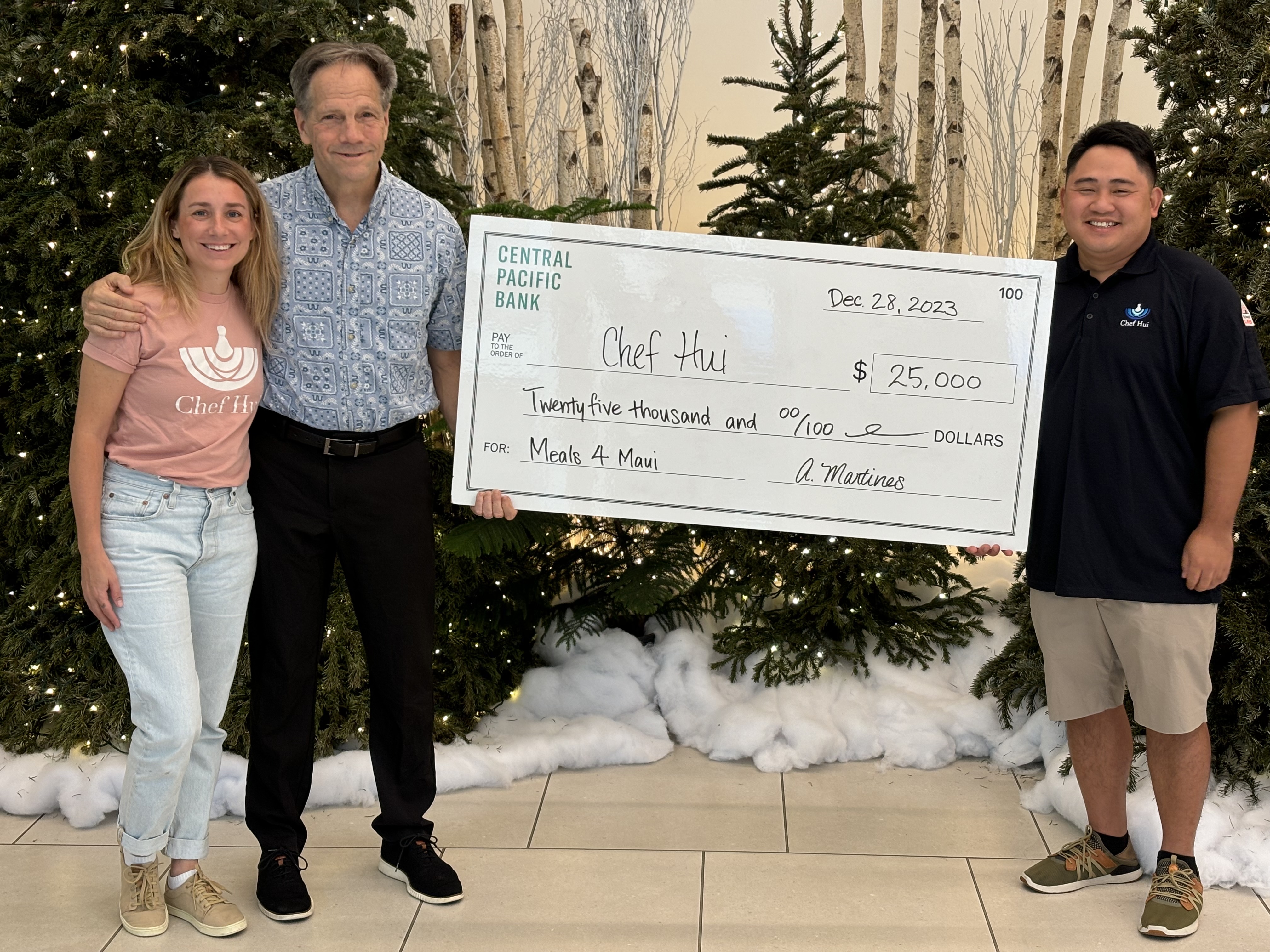 Three people stand indoors holding a large $25,000 Central Pacific Bank donation check for Chef Hui’s Meals 4 Maui program. The check presentation takes place in front of holiday decorations and evergreen trees, supporting meal relief efforts for Maui wildfire survivors.