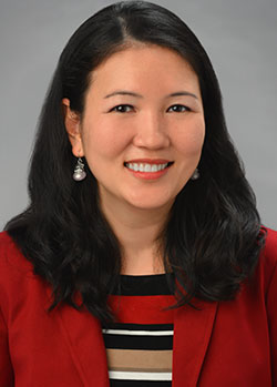 A professional headshot of a person wearing a red blazer over a striped top, photographed against a gray studio background.