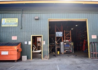 Exterior of a commercial warehouse with dark green siding, an open roll-up garage door revealing shelving and equipment inside, a side entry door, and an orange dumpster positioned outside.