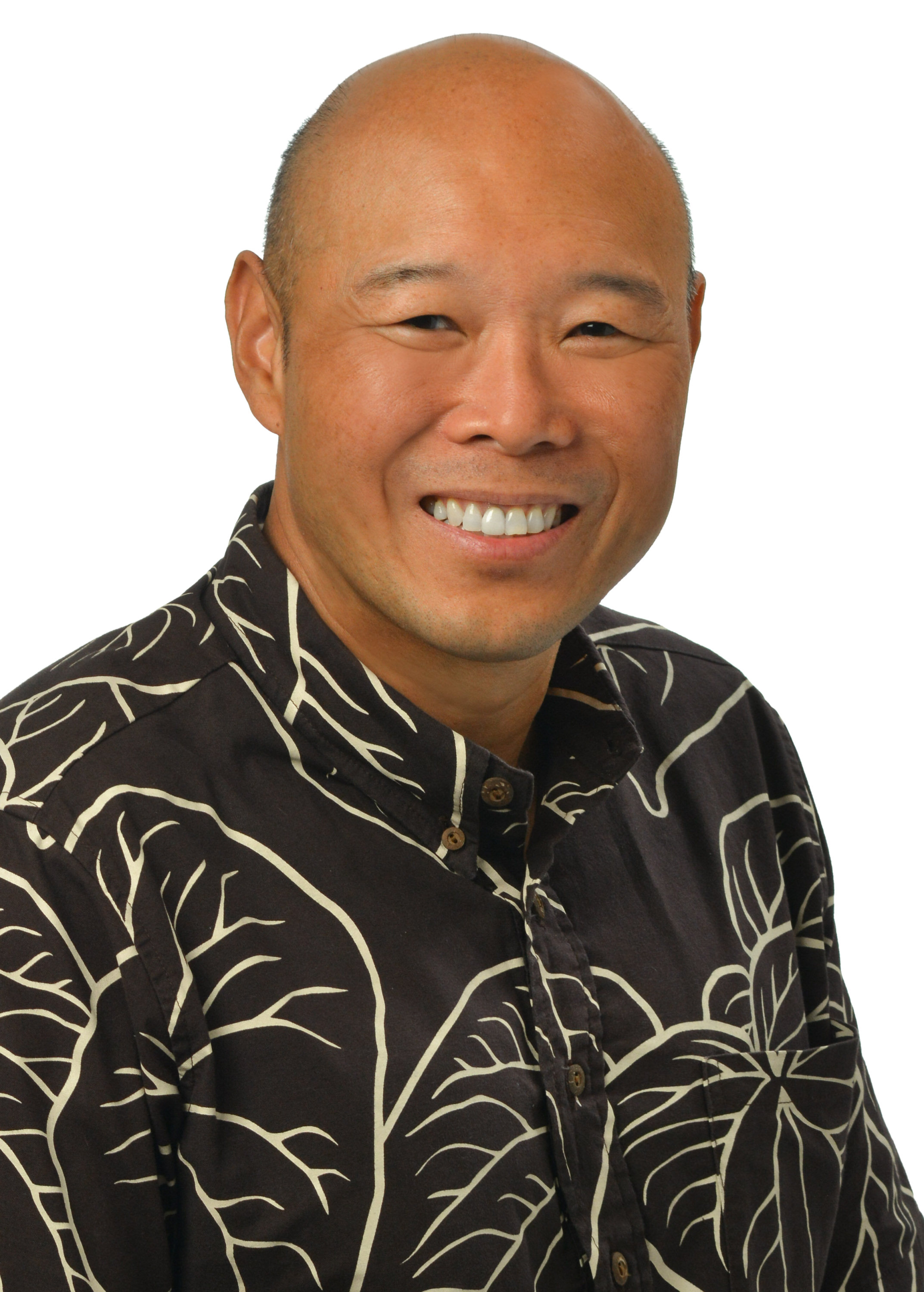 A professional headshot of a person with a shaved head wearing a black aloha shirt with a white leaf pattern, photographed against a light gray background.