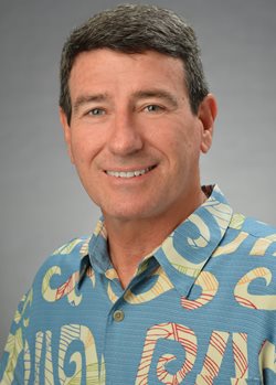 A professional headshot of a person wearing a blue patterned Hawaiian shirt, photographed against a gray studio background.