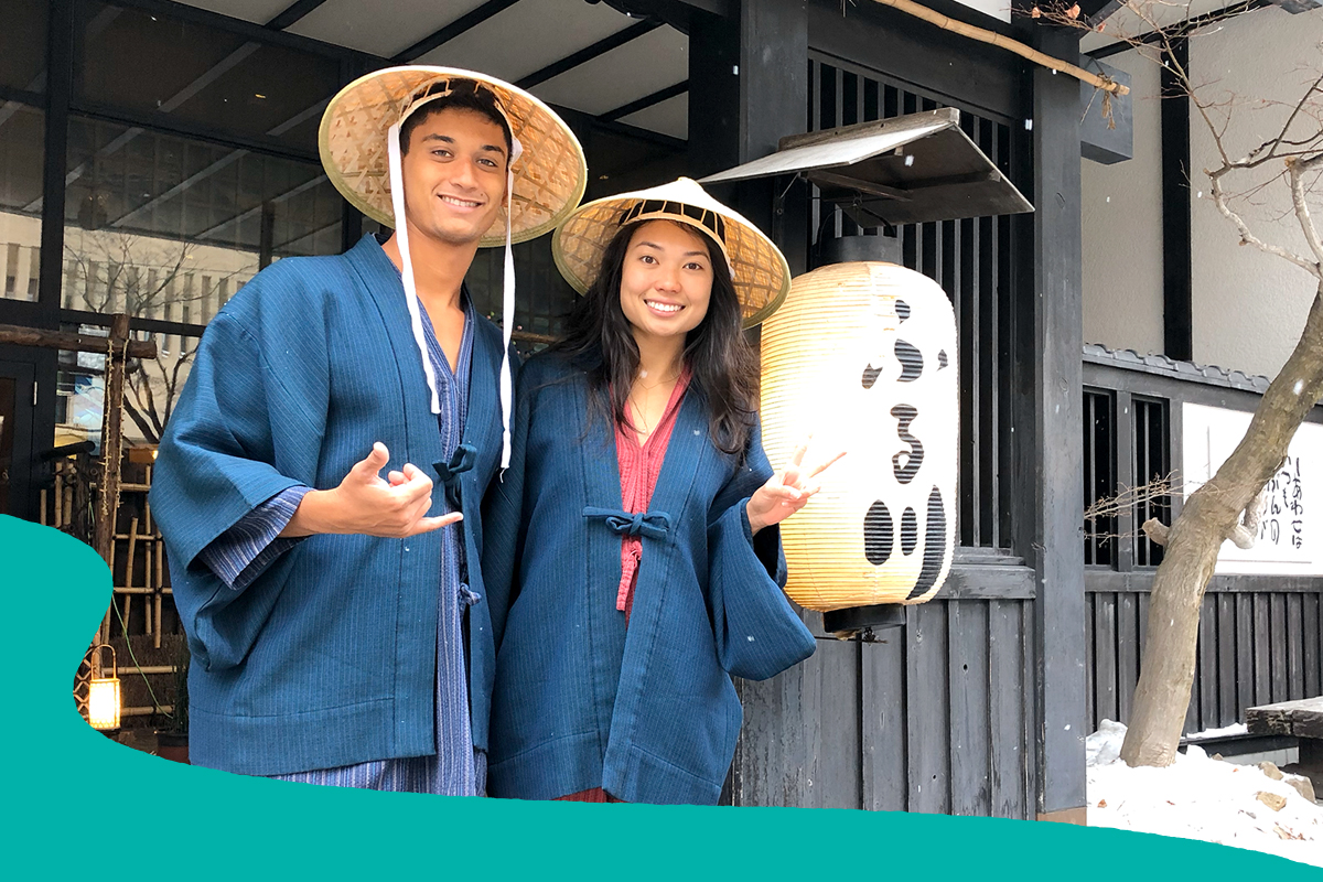 Two people wearing traditional Japanese robes and woven hats smile outside a Japanese-style building, with one making a shaka gesture and the other a peace sign.