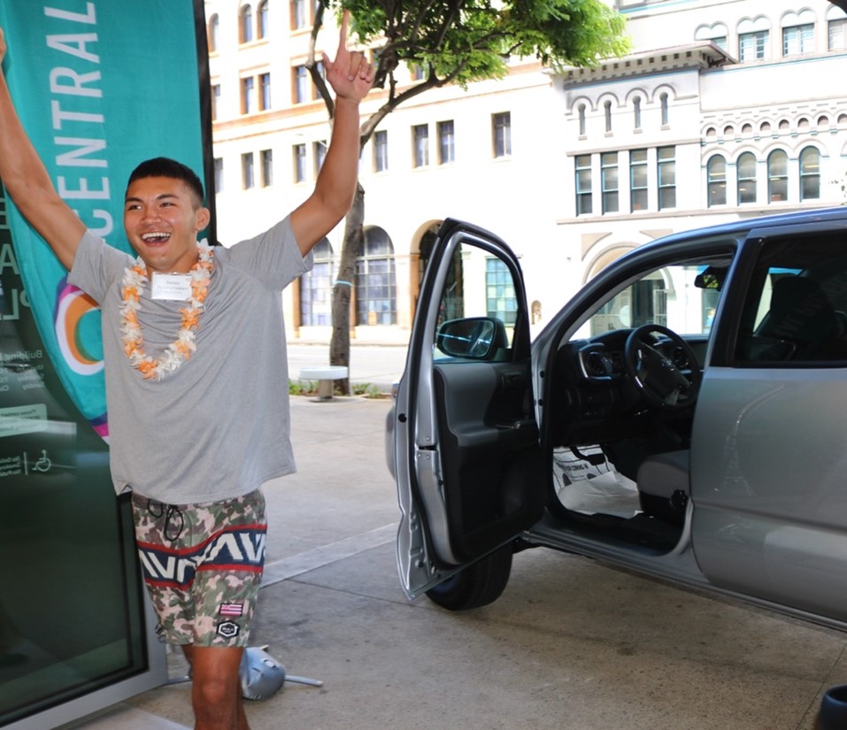 A person wearing a flower lei stands beside an open silver pickup truck outside a Central Pacific Bank branch, raising both arms during a vehicle giveaway celebration. Downtown buildings and bank signage are visible in the background.
