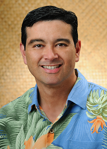 A professional headshot of a person wearing a blue floral Hawaiian shirt, photographed against a textured tan background.