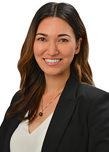 A professional headshot of a person wearing a black blazer over a white blouse and a pendant necklace, photographed against a light gray background.