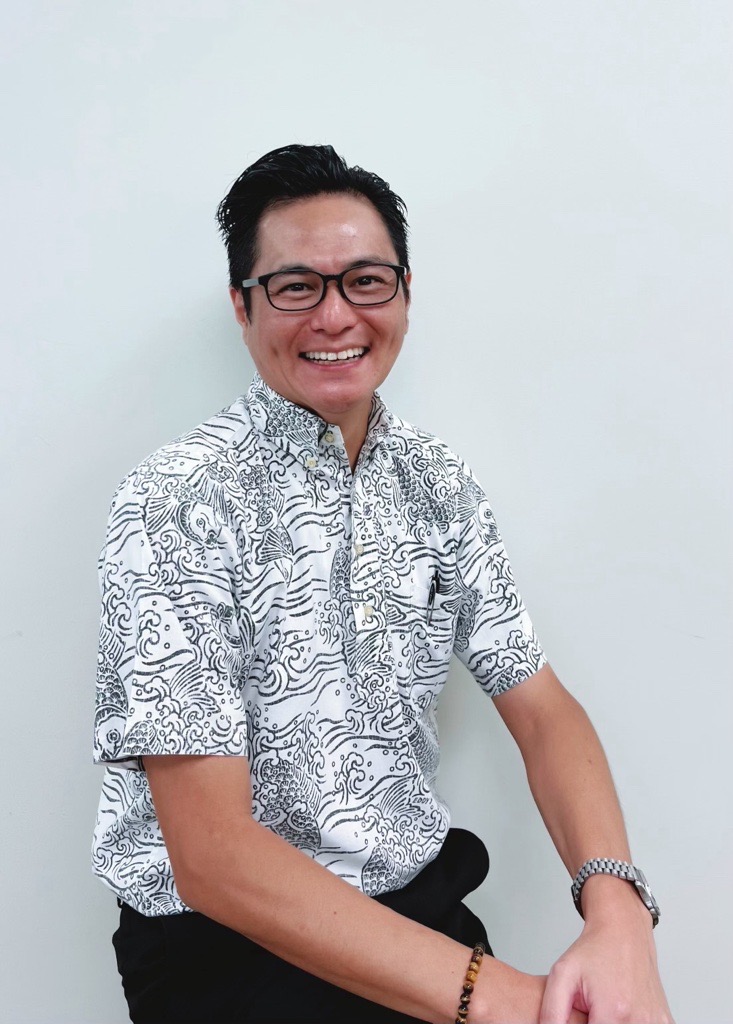 Smiling person wearing glasses and a patterned aloha shirt while seated against a plain background.