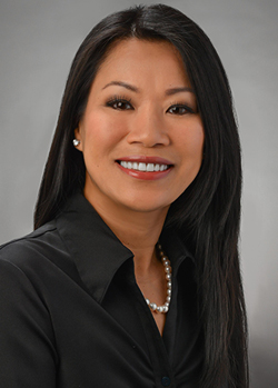 A professional headshot of a person wearing a black collared blouse and pearl necklace, photographed against a gray studio background.