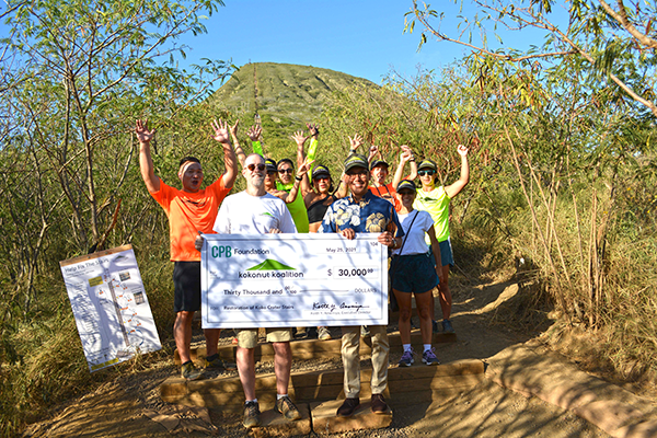 A group of community volunteers stand on a trail in front of a mountain landscape, holding a $30,000 CPB Foundation donation check for the Kokohead/Kokonut Koalition. Several participants raise their hands while posing outdoors among trees and vegetation.