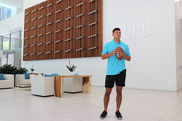 A person in athletic attire stands in the lobby of Central Pacific Bank holding a football. The bright, modern space features lounge seating, a decorative wood wall installation, and the letters “CPB” displayed on the wall in the background.