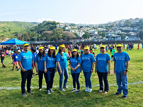  A group of volunteers wearing matching blue shirts and yellow caps pose for a photo on a grassy field during a community event, with participants and hillside homes visible in the background.