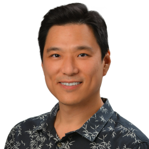 A professional headshot of a person wearing a dark aloha shirt with a light floral pattern, photographed against a light gray background.