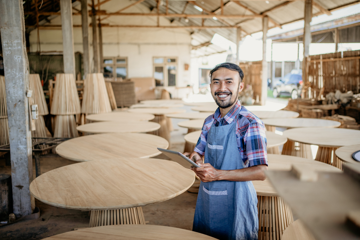 Furniture maker in an apron using a tablet inside a woodworking shop filled with unfinished wooden tables ready for production.