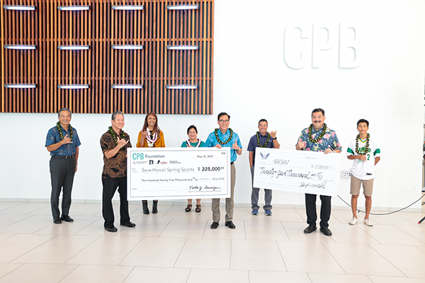 A group of eight people stand in the Central Pacific Bank lobby holding large donation checks for Save Hawaii's Sports. The recipients pose beneath the CPB logo and a decorative wood feature wall during a check presentation ceremony.