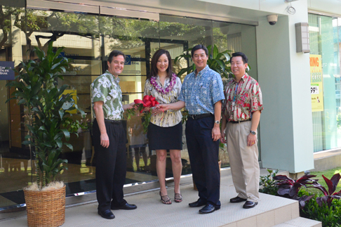 Four people stand outside a building entrance holding a large red ceremonial bow, posing in front of glass doors during an opening or celebration event.