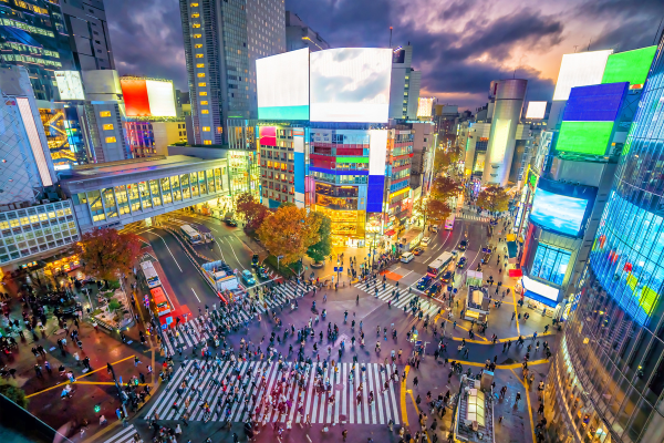 Elevated view of the Shibuya Scramble Crossing in Tokyo at dusk, with pedestrians crossing busy intersections surrounded by illuminated buildings and digital billboards.