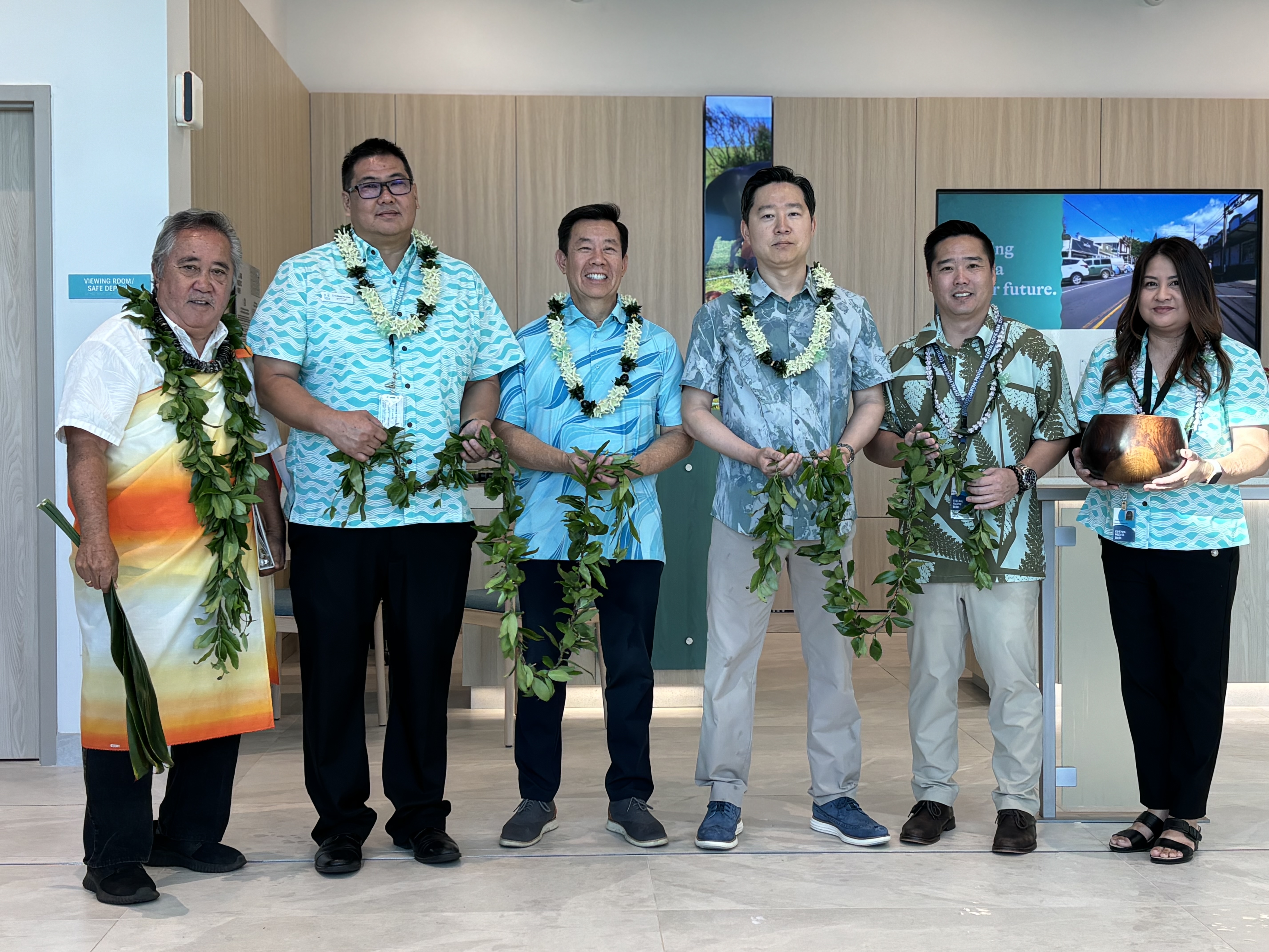 Six people wearing leis stand together in a modern bank lobby holding traditional greenery during a Hawaiian blessing or opening ceremony. The group poses in front of wood-paneled walls and digital displays inside a newly renovated branch.
