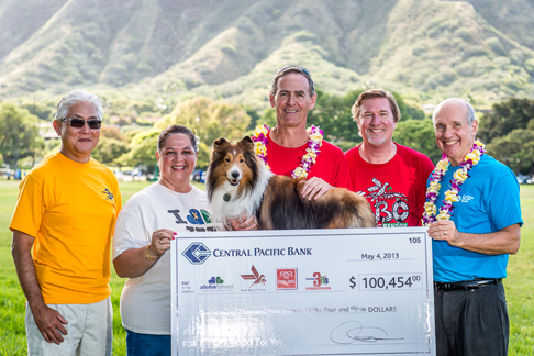 Five people stand outdoors with a Collie dog and a ceremonial donation check from Central Pacific Bank, with green mountains and a grassy field in the background.