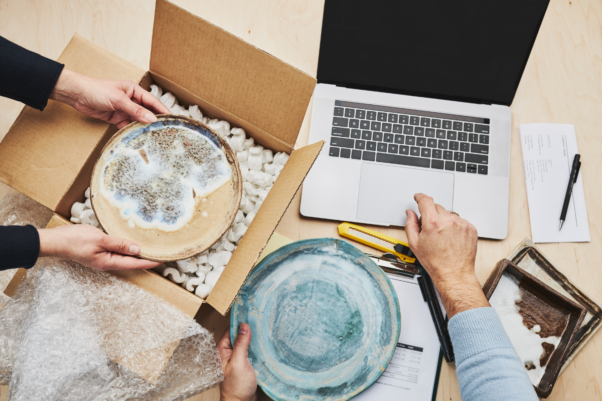 Overhead view of two people carefully packaging ceramic plates with protective packing materials next to a laptop and shipping supplies.