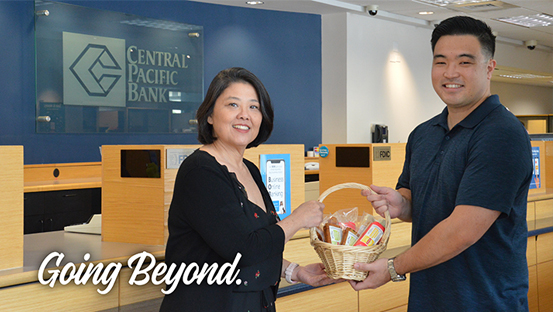 A person presents a gift basket to another person inside a Central Pacific Bank branch, with the bank logo displayed in the background and the words “Going Beyond.” shown in the lower corner.