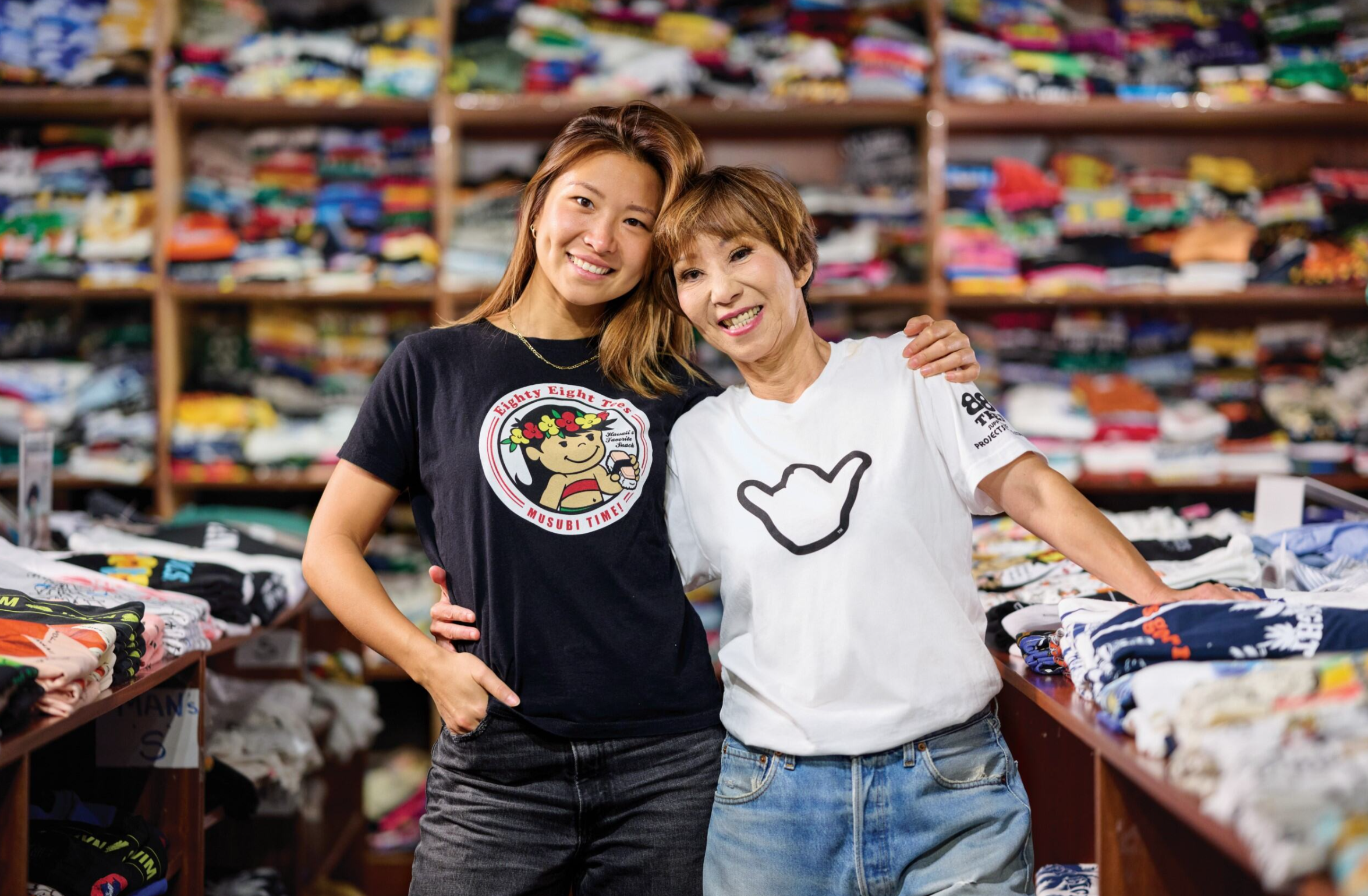 Two people posing together in a retail shop filled with neatly folded graphic T-shirts, smiling with their arms around each other.