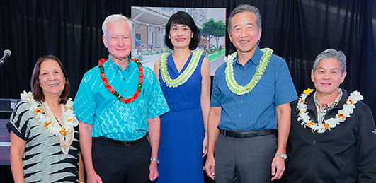 A group of five people wearing floral leis pose together at an indoor event, standing in front of a black backdrop with a building rendering displayed behind them. The group is dressed in business and aloha attire and gathered for a community or organizational gathering.