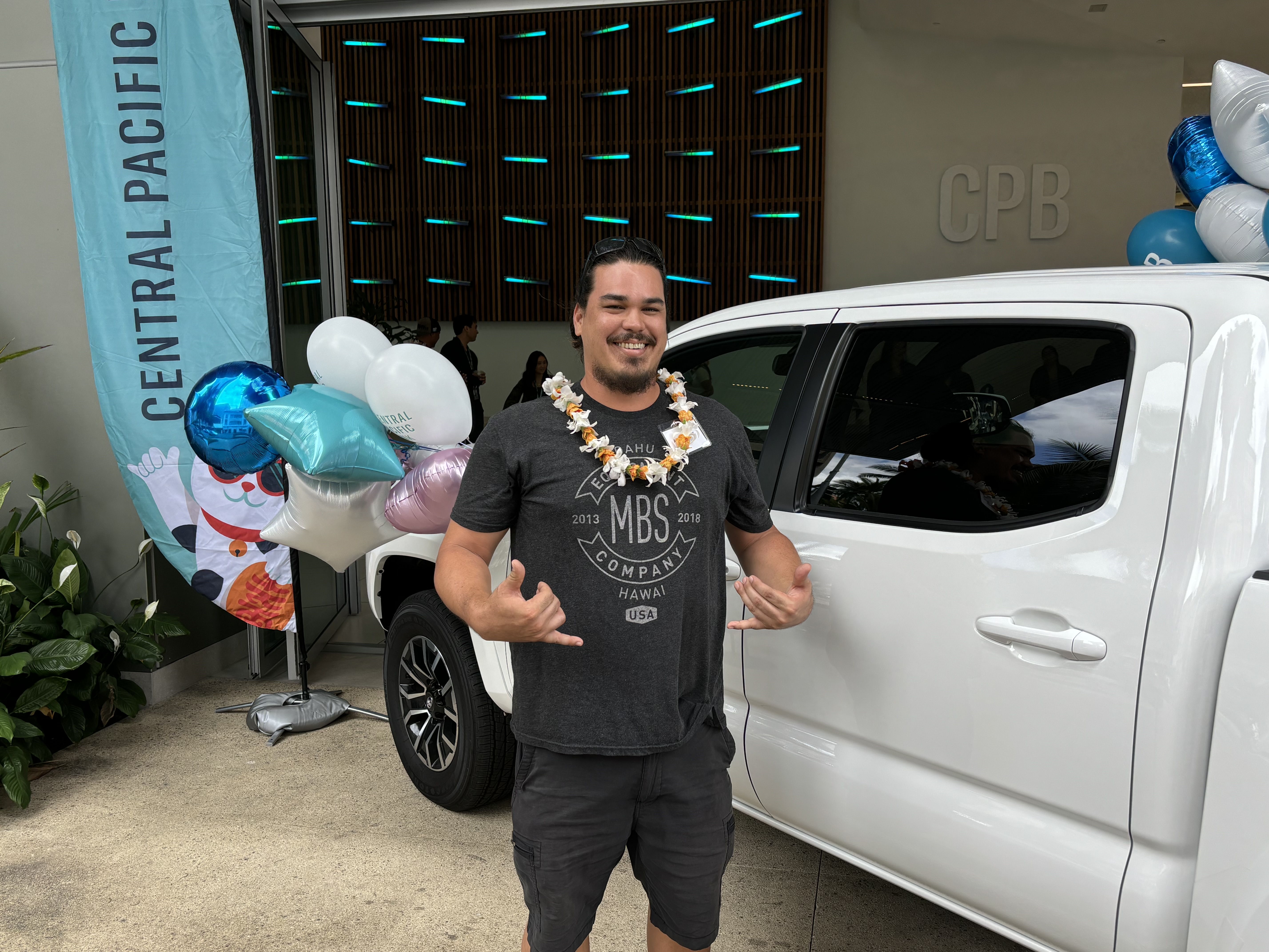 A person wearing a flower lei stands beside a white Toyota Tacoma pickup truck outside a Central Pacific Bank branch decorated with balloons and promotional signage. The person makes shaka gestures while posing next to the truck during a vehicle giveaway celebration.