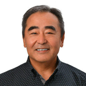 A professional headshot of a person wearing a dark patterned button-down shirt, photographed against a light gray background.