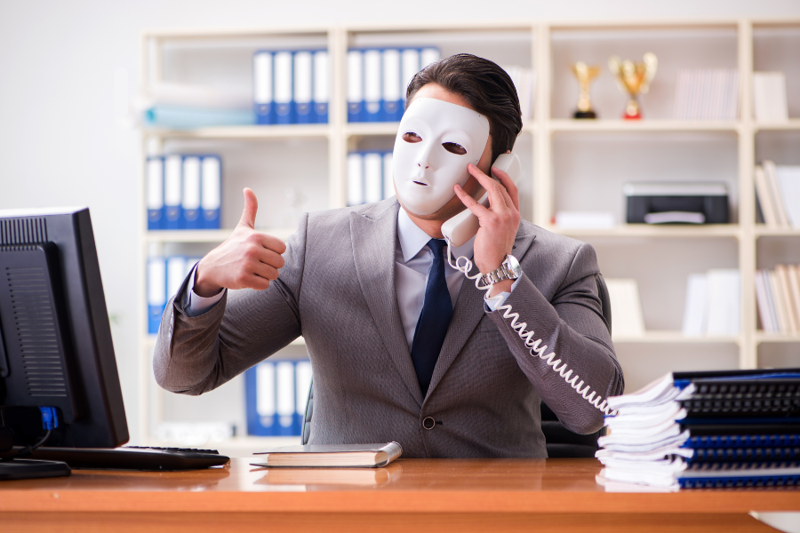 Person in business attire wearing a white mask while speaking on a desk phone in an office, illustrating a scammer impersonating a trusted caller.