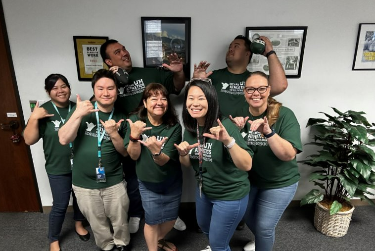 Seven coworkers in the HR department wearing matching green shirts pose together in an office, smiling and making shaka hand signs.