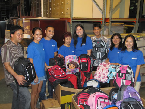 Volunteers standing in a warehouse surrounded by colorful backpacks collected for a back-to-school donation program.