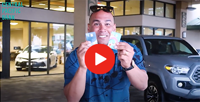 A person stands outside a Central Pacific Bank branch holding two bank cards, with parked vehicles and the bank entrance visible in the background. A video play button overlay appears in the center of the image.