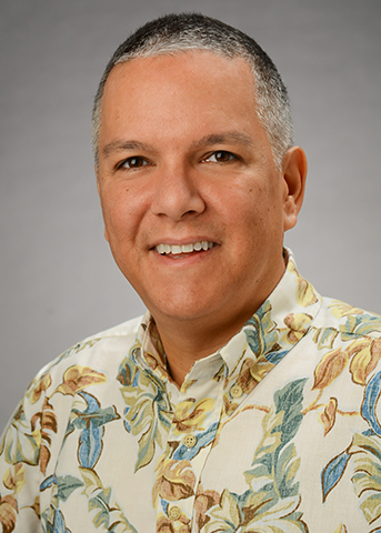A professional headshot of a person wearing a cream-colored floral Hawaiian shirt, photographed against a gray studio background.