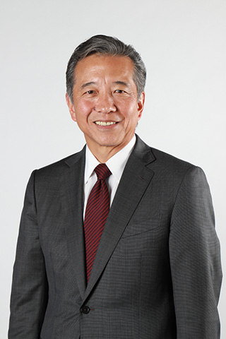 A professional business portrait of a person wearing a dark gray suit, white dress shirt, and red striped tie, standing against a light gray studio background.