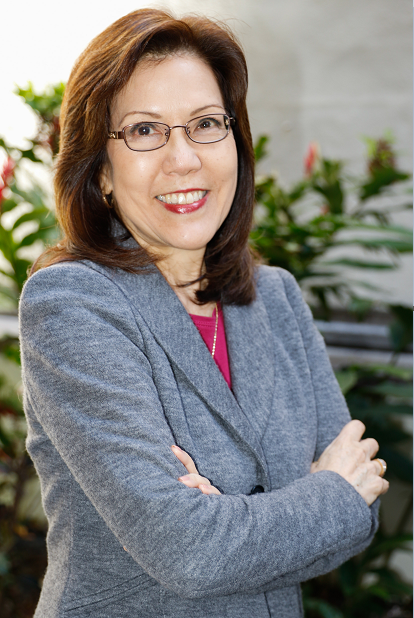 A professional portrait of a person wearing glasses and a gray blazer over a pink top, standing with arms crossed in an office setting with green plants in the background.