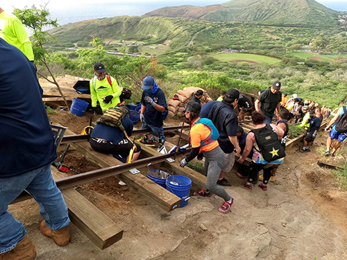 A group of volunteers works together on a mountainside trail improvement project, carrying and installing materials along a steep slope. The image overlooks a lush green valley and surrounding ridgelines, highlighting the scale of the outdoor restoration effort.