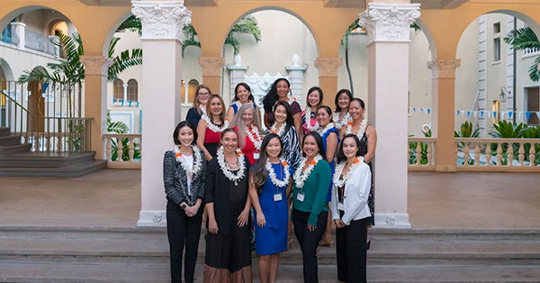A group of professionals wearing floral leis pose for a group photo on the steps of an open-air courtyard with decorative arches and columns. The group stands in front of a tropical-inspired building entrance surrounded by palm trees and architectural details.