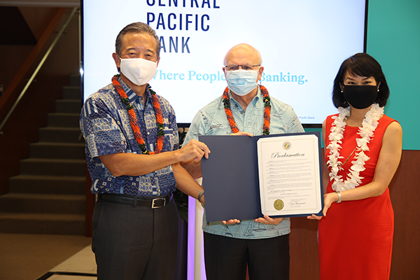 Three people wearing face masks and leis hold a framed proclamation during a recognition ceremony at Central Pacific Bank. The presentation takes place in front of a screen displaying the Central Pacific Bank logo and the slogan, “Where People Like Banking.”