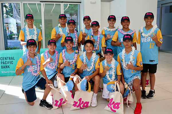 A youth baseball team in matching blue and yellow uniforms poses for a group photo inside the Central Pacific Bank lobby. Team members wear leis, hold tote bags and caps, and make shaka gestures while standing in front of large windows and bank signage.