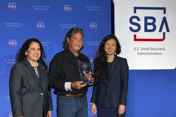 Three people pose with an award at a U.S. Small Business Administration (SBA) event, standing in front of SBA-branded displays. One person holds a recognition plaque while the group gathers for a photo during an SBA awards ceremony.